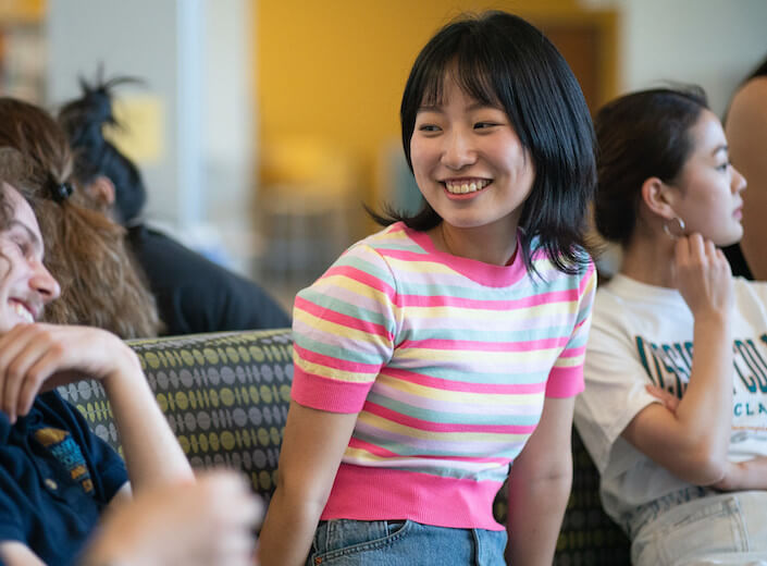 Young Asian woman in short-sleeved pink and yellow striped sweater top, black bob, and fair skin leans forward and smiles in SEC building.
