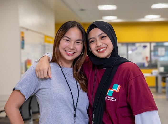Two young women pose together in the Academic Success Center. One wears a hojab, the other is East Asian.
