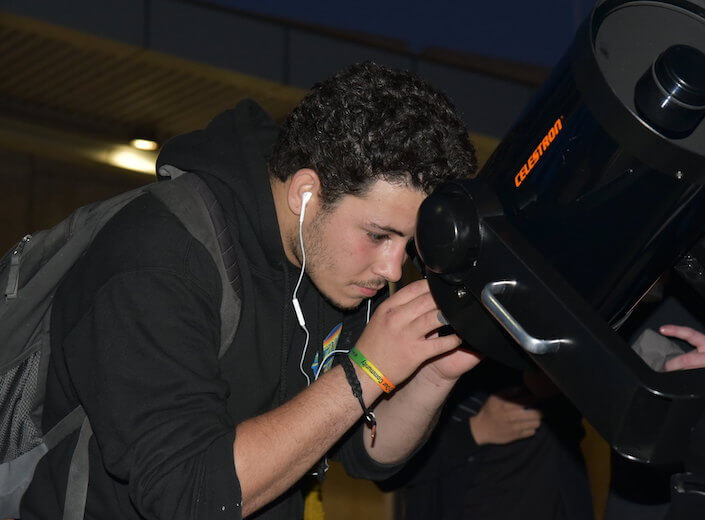 A young man with curly dark hair, a dark blue hoodie, and earbuds in looks into the viewfinder of a large telescope.