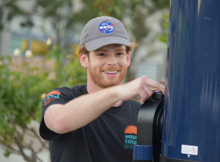 Astronomy lab. A young man with red hair and a NASA baseball hat adjusts a telescope for a Astronomy event on campus. He is smiling.