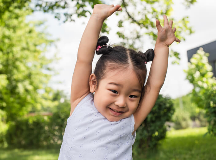 Asian girl toddler smiles and lifts her arms above her head.