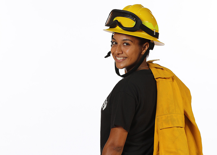 Latinx female student poses with a firefighter helmet and coat slung over her shoulder. She wears a black tshirt.
