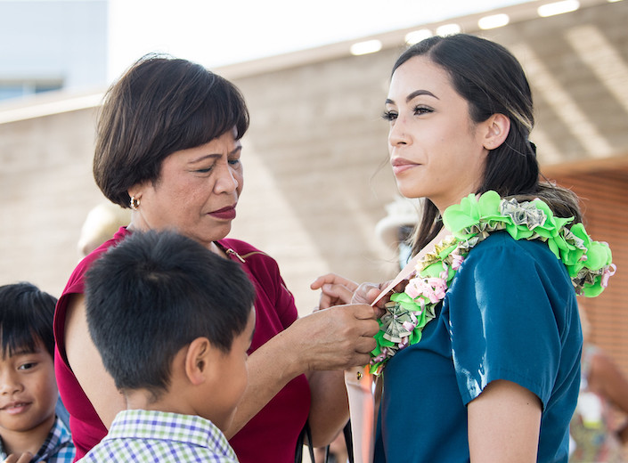 Young Filipina-American RN graduate wears her blue scrubs and stands as her mother "pins" her in a ceremony. She wears a flower leigh and has long, straight black hair.