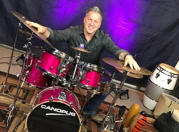 Brian Andres at his drum kit. He is a man with light skin and grey hair. His hands are outstretched on top of his drum kit, resting on gold symbols.