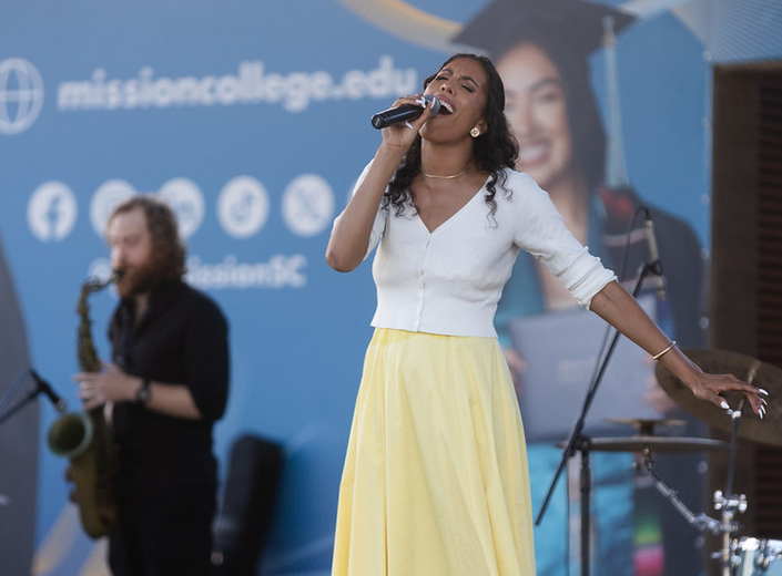 Female singer with long curly black hair and a long-sleeved shirt belts out a song into a microphone.