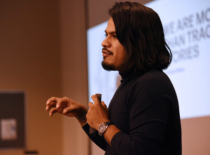 Yosimar Reyes is pictured in profile as he delivers his presentation at the Black and Brown Summit. He has black hair to his shoulders, a black turtleneck, and black facial hair.