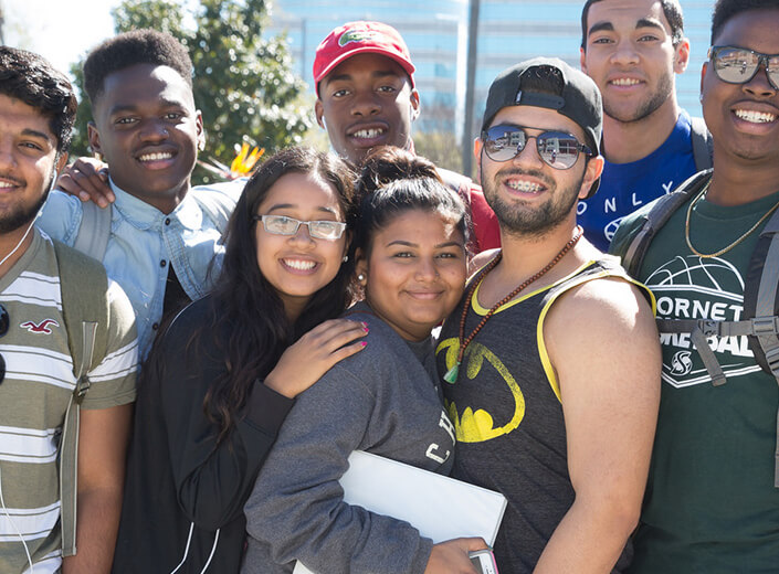 Group of students from various ethnic backgrounds and genders pose together in a group outside on a college campus.