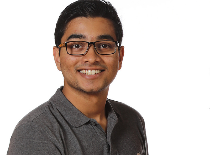 Male college student with light brown skin, black hair, a grey polo shirt, and glasses smiles.