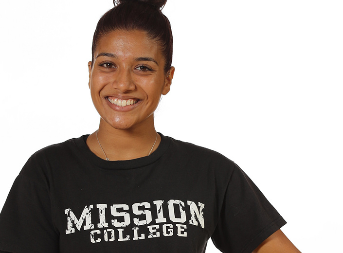 Young woman with a black Mission College Tshirt poses for camera. She has dark hair pulled back in a bun and a big smile. 