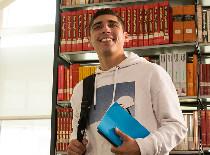 Latino college student wears a white hoodie and holds a handful of books in his college library. He stands in front of a shelf of books. His hair is short and black and he has a backpack slung over one shoulder. He is smiling.