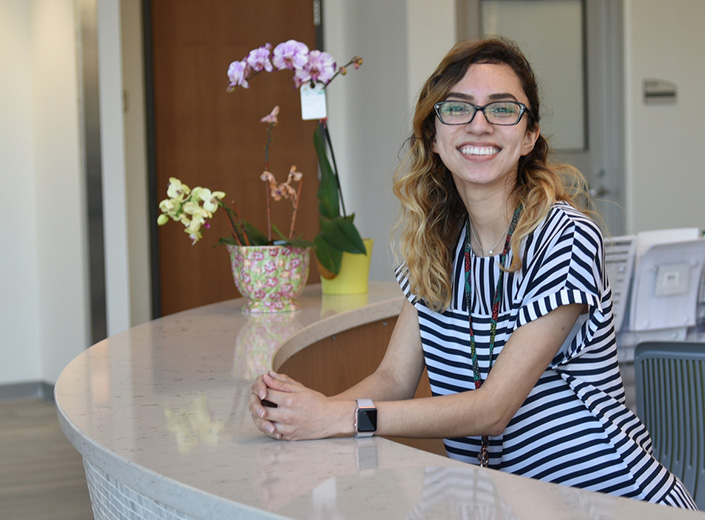 Young woman with striped tshirt leans on her elbows on a service counter/desk. She has light brown wavy hair and light brown skin. She wears classes.