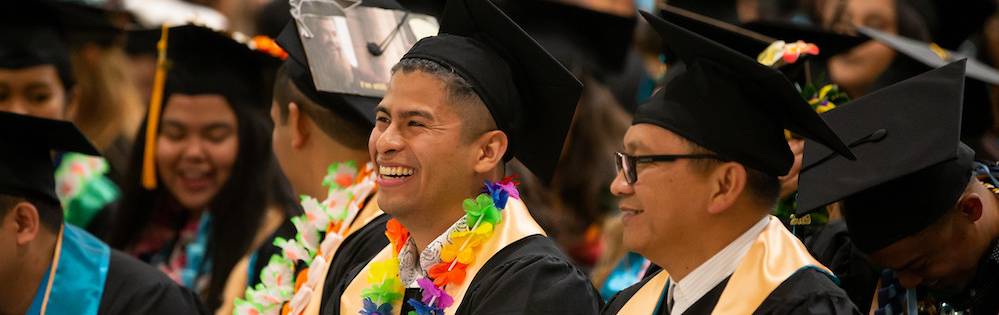 Two young Latino men in graduation regalia in the audience of the commencement ceremony of their community college.