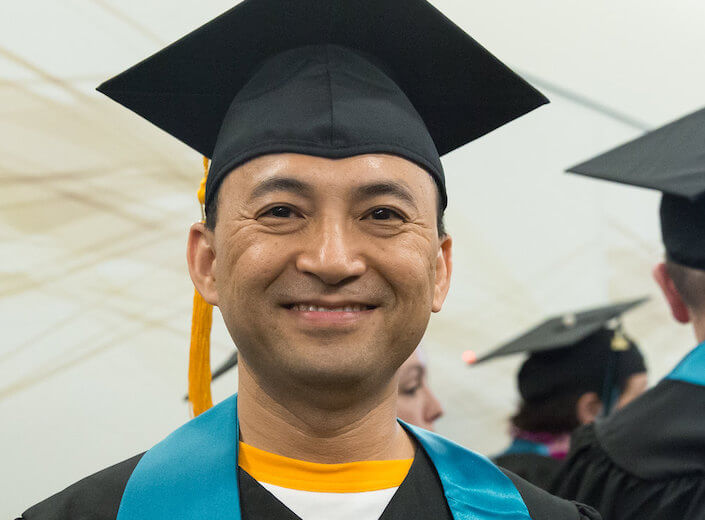 MIddle-aged Latino man in cap and gown smiles.