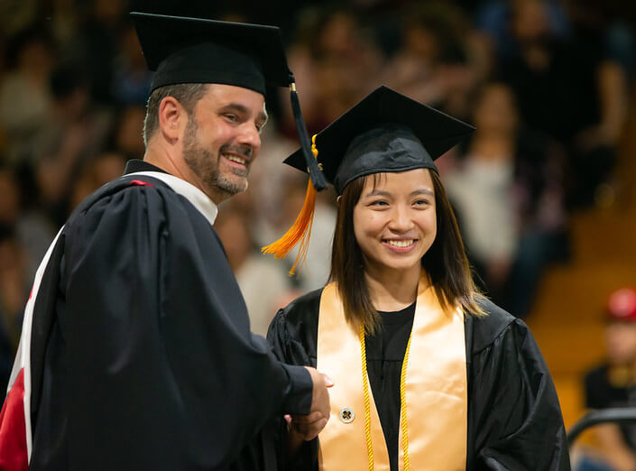 A smiling Asian-American female college student wears her commencement regalia and shakes hands with the college president, a white man with short brown hair (also wearing graduation regalia.) 