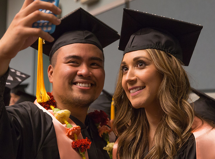 Male and female community college graduates take a selfie in their black cap and gown with colorful leighs around their neck.
