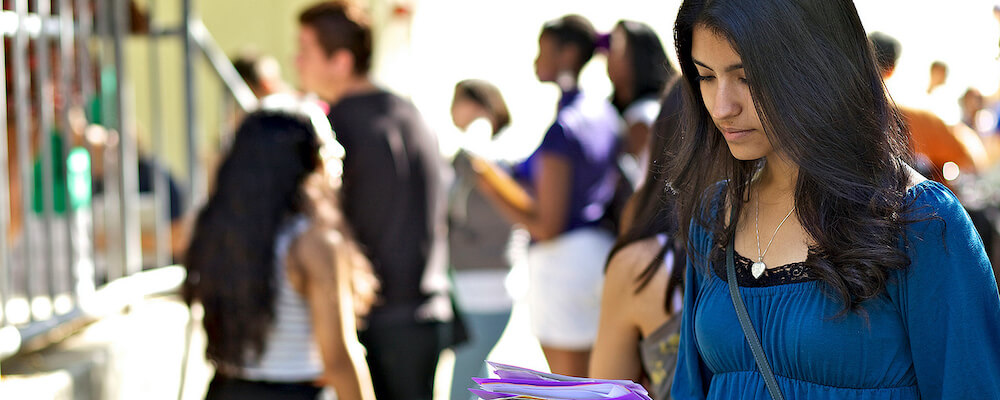 Young woman with long black hair looks down at a brochure in her hands as students behind her congregate in small groups.