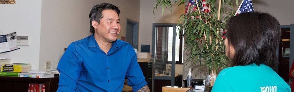 Man behind desk A man in a blue button-down shirt stands behind a desk in a college office and smiles toward a student who is pictured from behind. She has shoulder-length black hair.