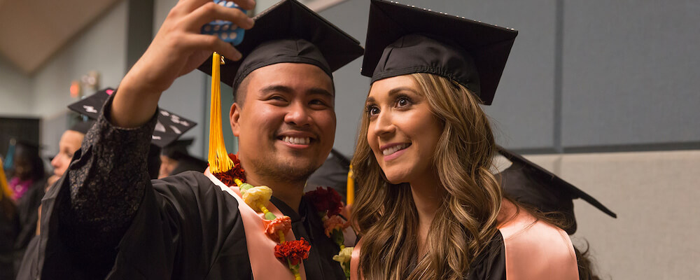 A male and female student take a selfie together at commencement. They are wearing graduation regalia decorated with floral leis. 