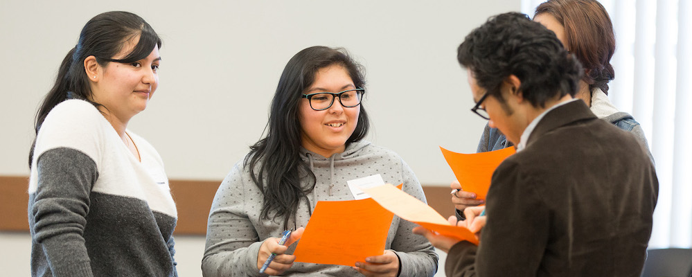 Puente Three students of LatinX descent hold bright folders in a classroom.