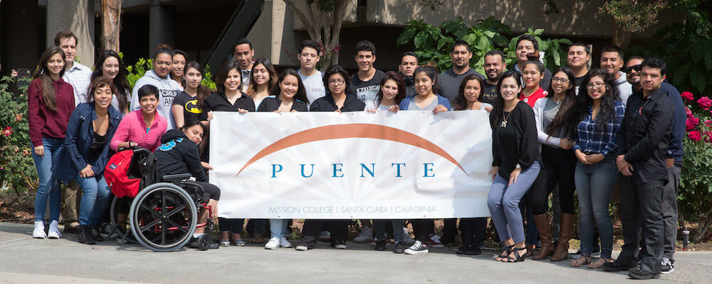 A group of students pose in a group outside and hold a sign that reads "Puente".
