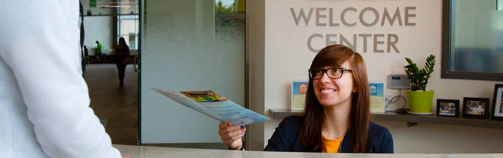 Young woman with glasses and straight brown hair hands a brochure to a student over the desk at the Welcome Center.