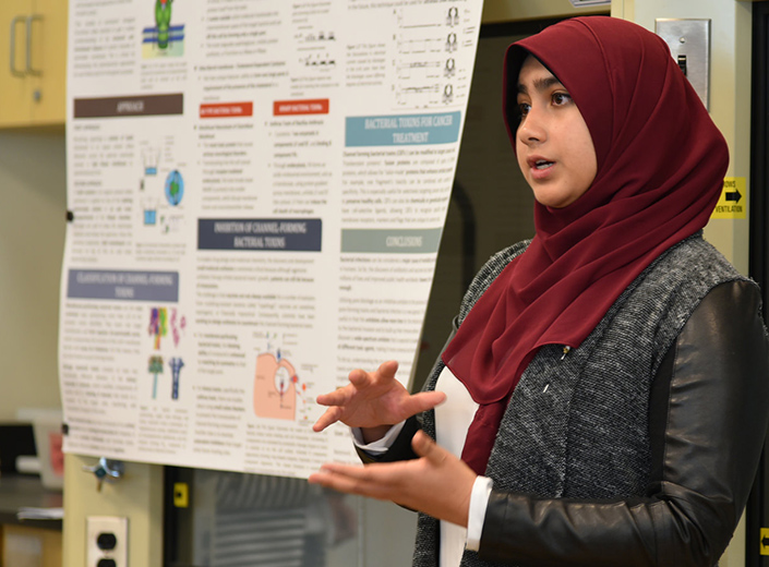 Young woman in red hijab presents a science project. Her ideas are presented on a poster board behind her.