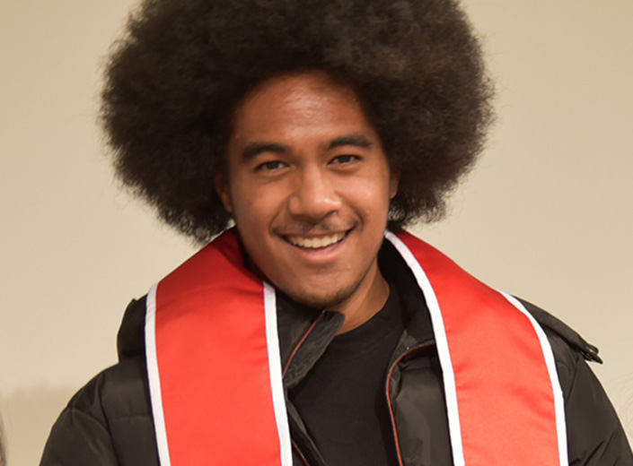 Young man with natural grown-out curly hair and moustache wears a bright organe "MESA" sash at a commencement ceremony.