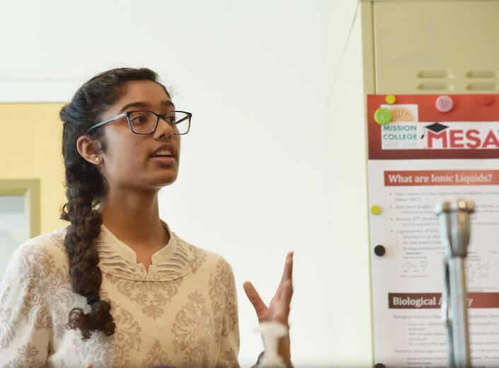 Young woman of South Asian descent wears glasses and explains a science project during a MESA event.