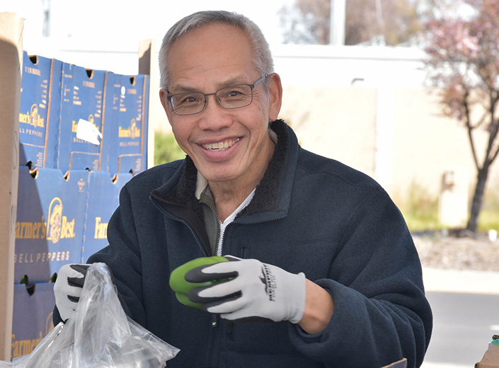 Second Harvest Food Bank. An older man of Asian-American descent wears gloves and puts a pepper in a plastic bag. He wears a blue sweater and wire-framed glasses.