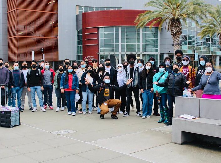 A group of students (male, female, students of color) are posed in a group outside on campus in casual clothes on their way to a college tour of SJSU.
