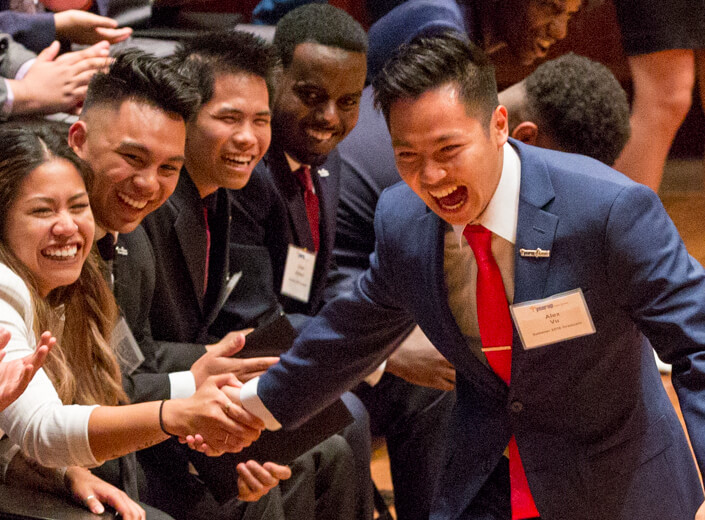 Asian-American male grad in a blue suit runs by front row of peers in a Year Up graduation ceremony.